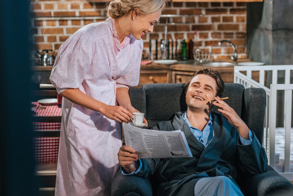 A woman in a pink robe hands a man in a dark robe a cup of coffee as he sits in a chair, holding a newspaper and a cigarette, in a kitchen setting.