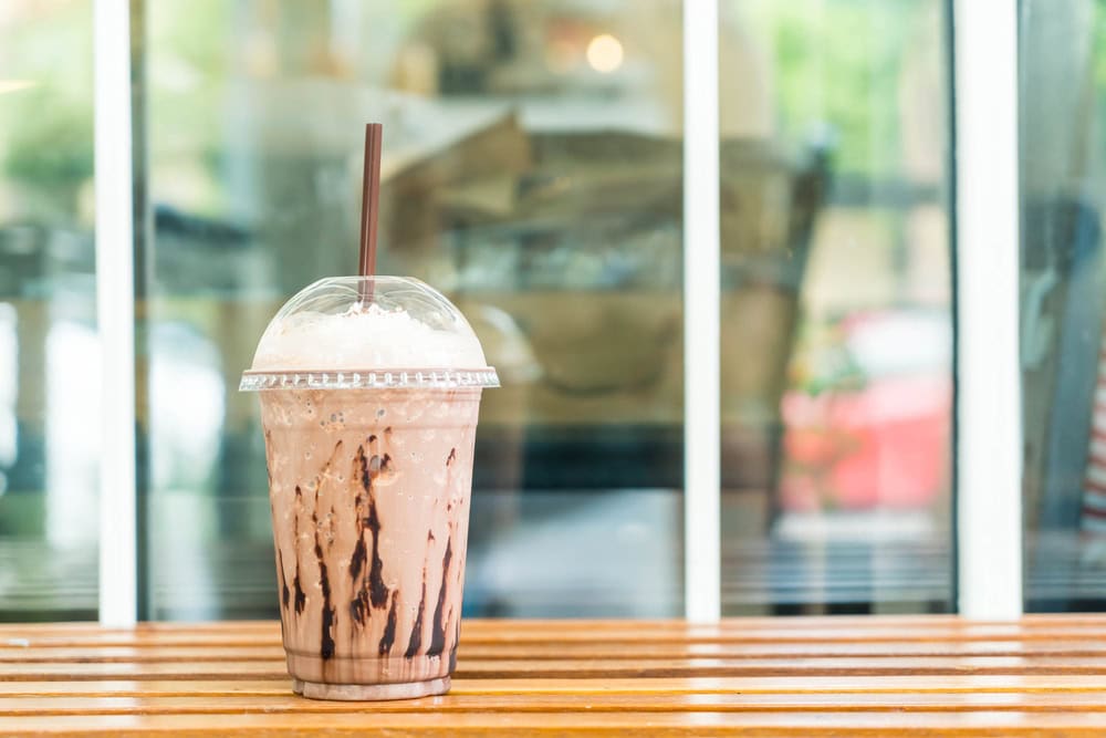Iced chocolate drink with whipped cream and a straw in a clear plastic cup, placed on a wooden table in front of a window.