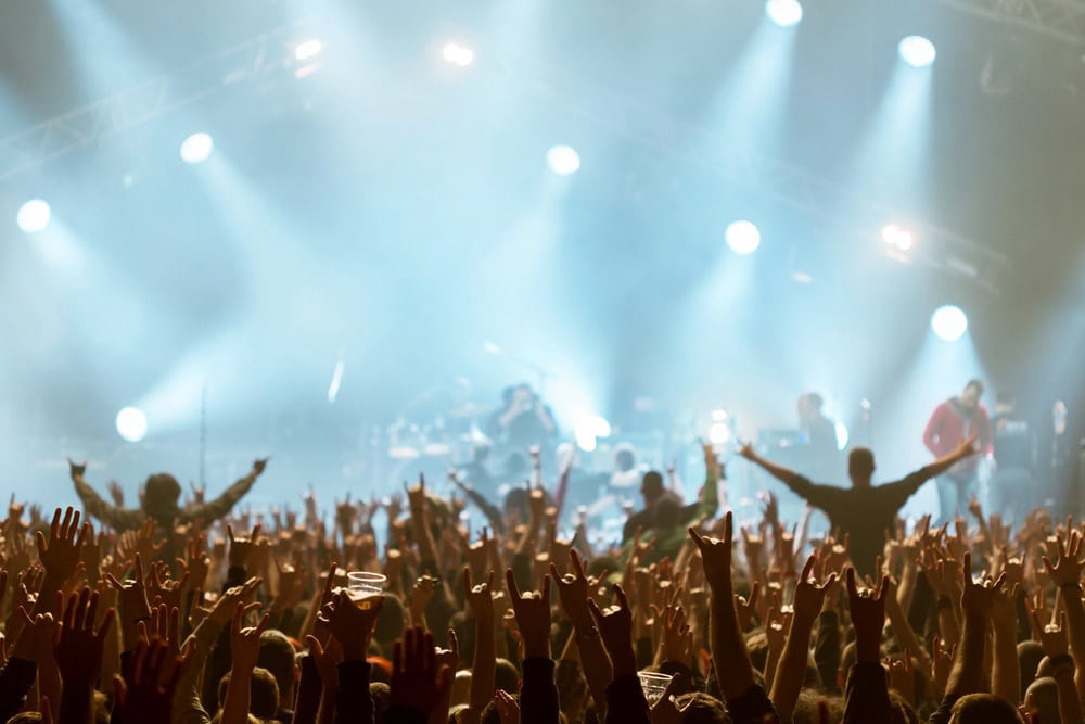 A crowd at a concert with hands raised, facing a brightly lit stage where a band is performing under blue and white stage lights.