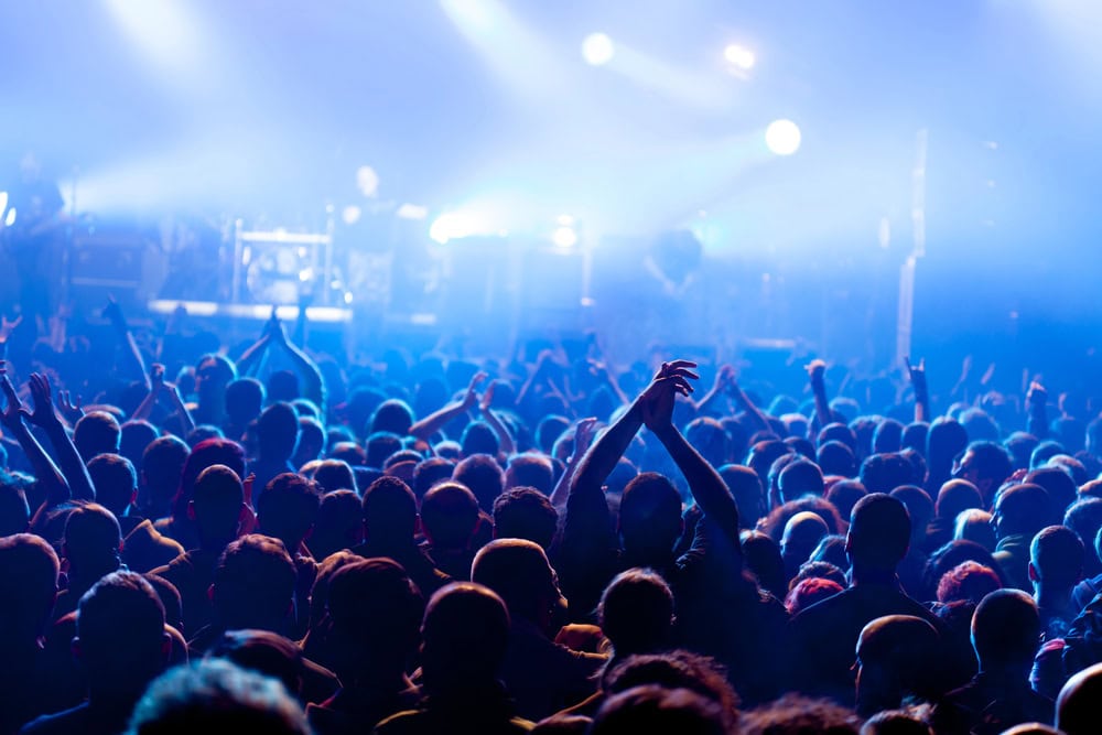 A large crowd faces a brightly lit stage at a concert, with raised hands and blue lighting filling the scene.