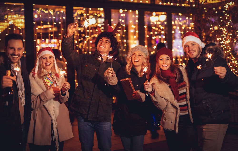 Six people dressed in winter clothing and festive hats stand outdoors at night, holding sparklers and smiling in front of holiday lights.