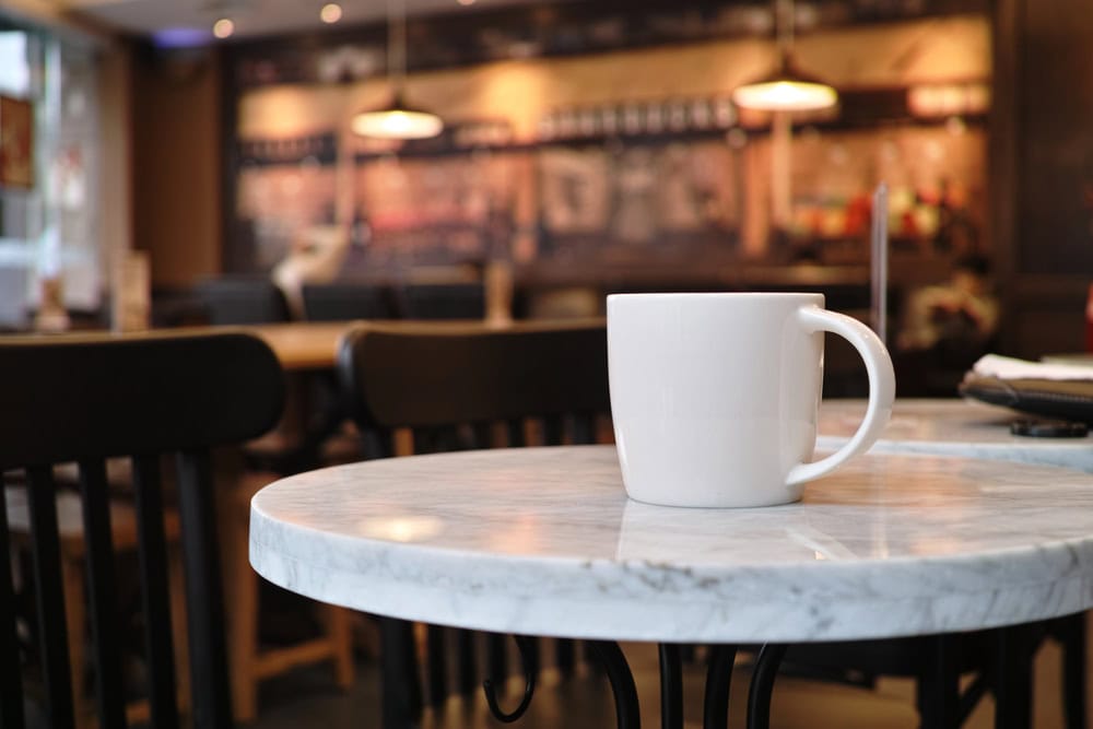 A white coffee mug sits on a round marble table in a café with wooden chairs and warm lighting in the background.