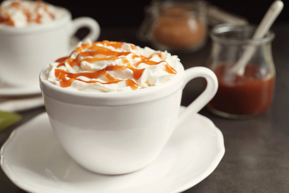 A white cup filled with a whipped cream-topped drink, drizzled with caramel sauce, sits on a saucer; jars of caramel sauce are in the background.