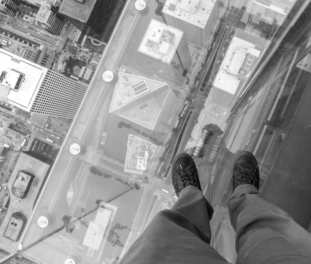 A person stands on a glass floor high above a city, looking down at buildings and streets far below.