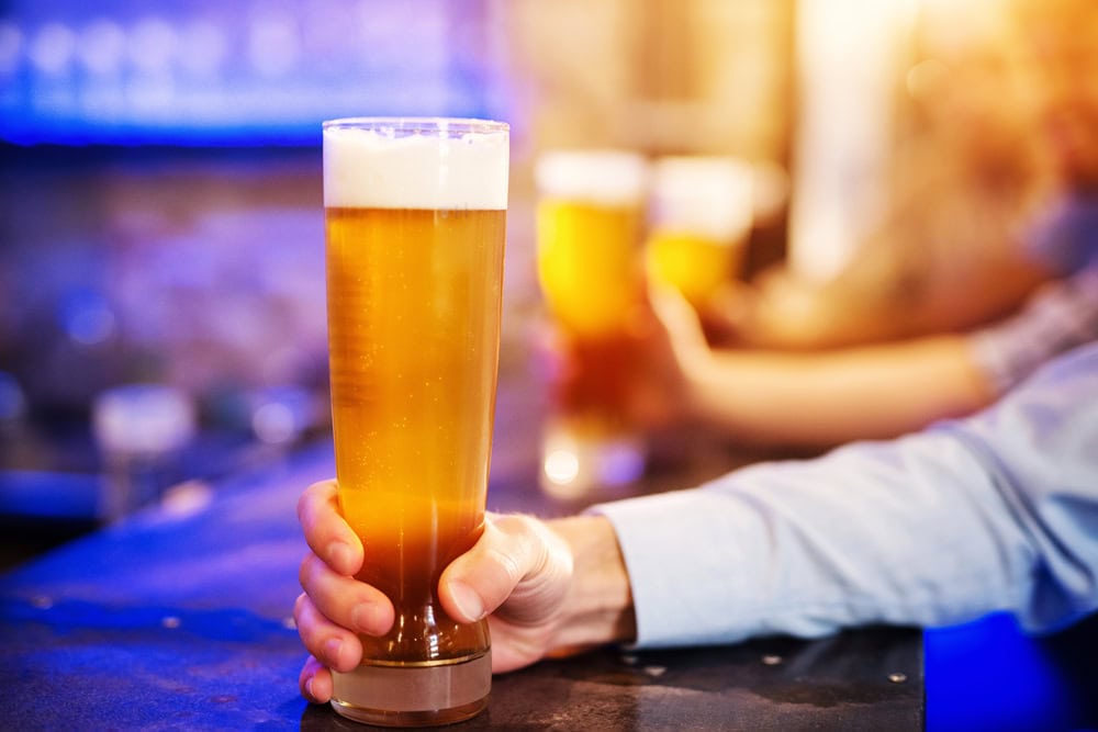 A person holding a tall glass of beer with foam on top at a bar, with other glasses of beer visible in the background.