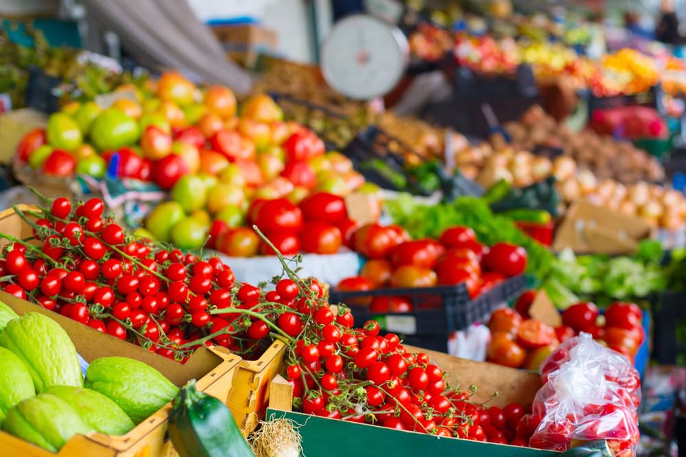 Fresh fruits and vegetables, including tomatoes, apples, lettuce, and squash, displayed in crates and bins at an outdoor market.
