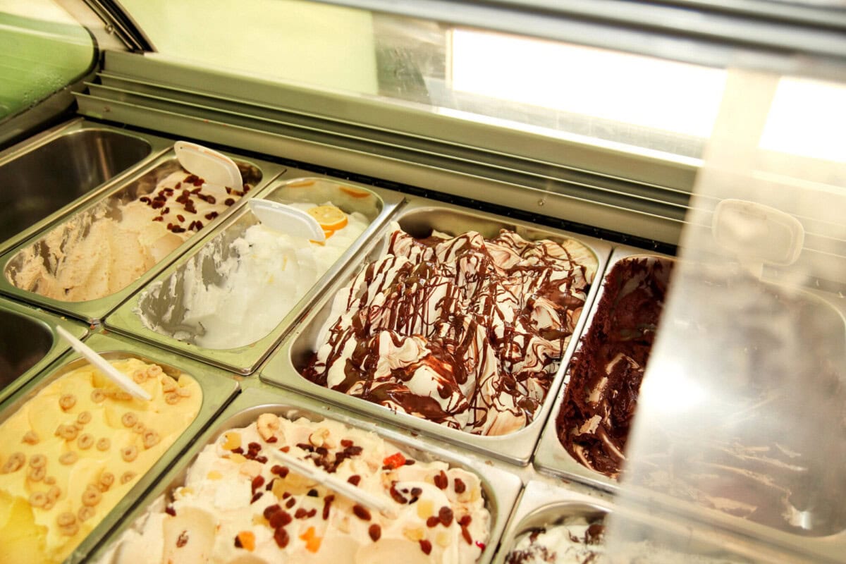 Display case with various flavors of gelato, including vanilla, chocolate, and fruit varieties, some topped with chocolate drizzle or fruit pieces.