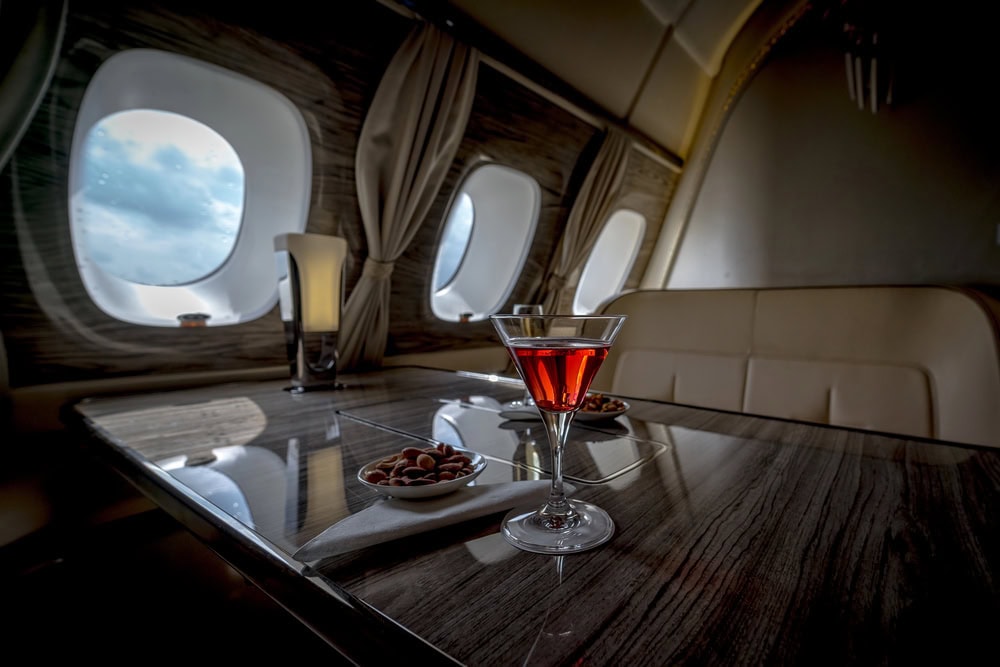 A glass of red cocktail and bowls of nuts on a table inside a private jet with large windows and beige seating.