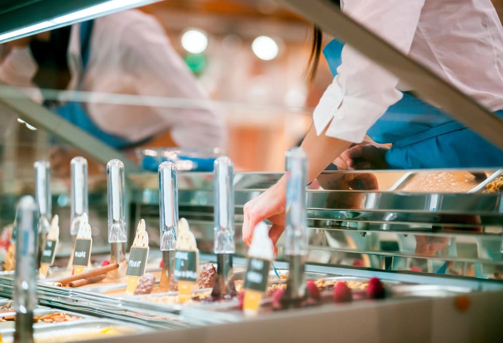 A person in a blue apron serves gelato behind a glass display case with various flavors and metal scoops visible.