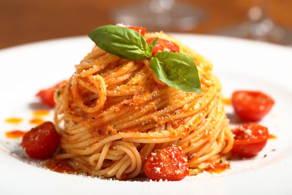 A serving of spaghetti with tomato sauce, cherry tomatoes, grated cheese, and fresh basil leaves on a white plate.