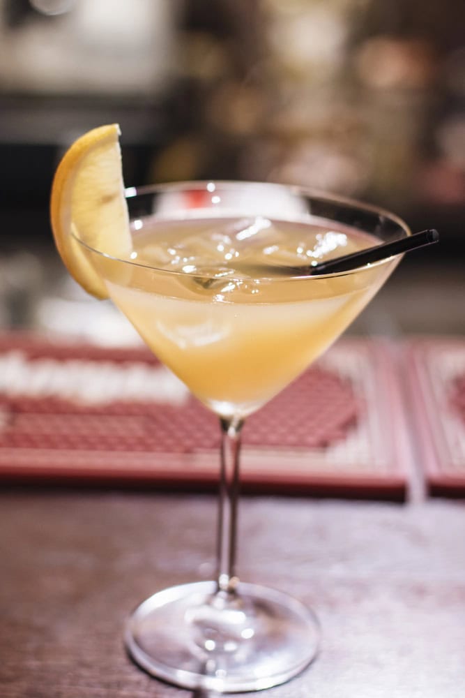 A cocktail served in a martini glass with ice, a lemon wedge garnish, and a black straw on a bar counter.