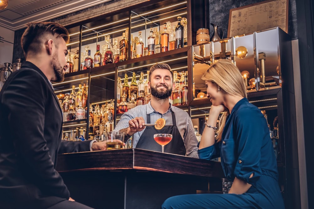 A bartender garnishes a cocktail for two seated patrons at a bar with shelves of liquor bottles in the background.