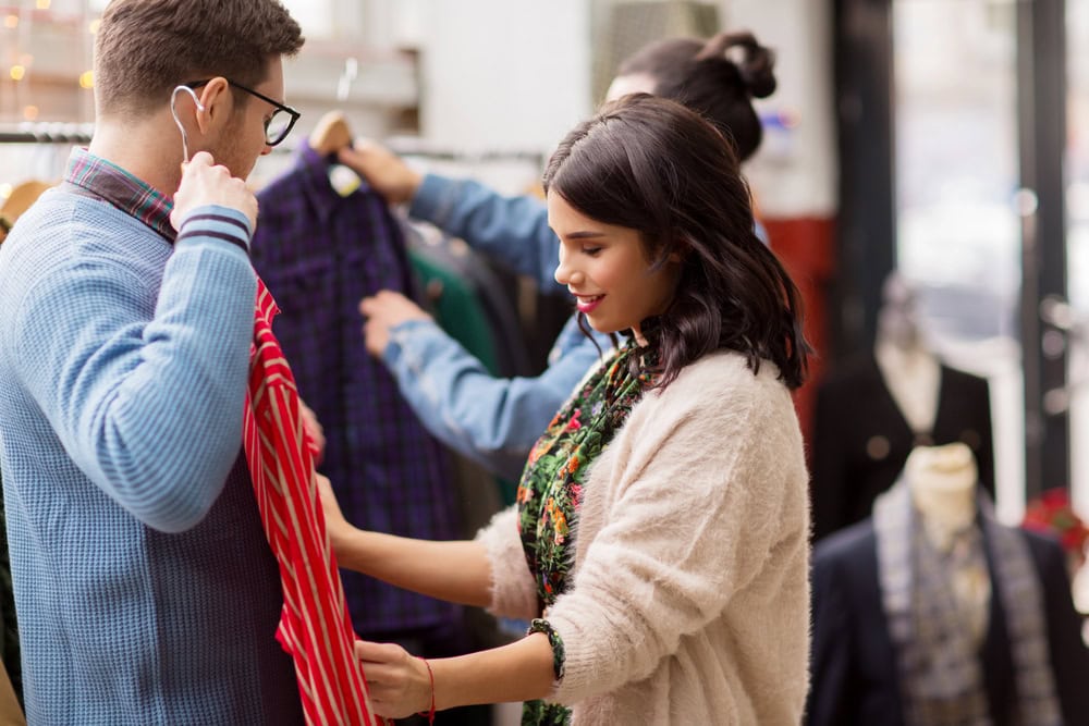 A woman helps a man try on a red striped shirt in a clothing store, with clothes and mannequins visible in the background.
