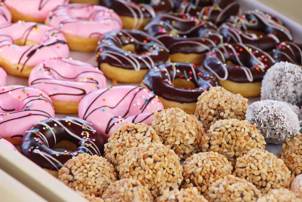 A display of pink and chocolate-glazed donuts with sprinkles, and assorted round pastries coated in chopped nuts and coconut.