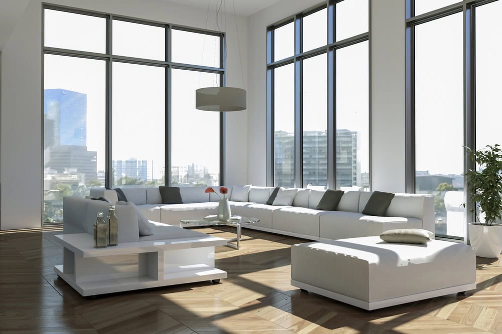Modern living room with large windows, white sectional sofa, a low coffee table, potted plant, and city buildings visible outside.