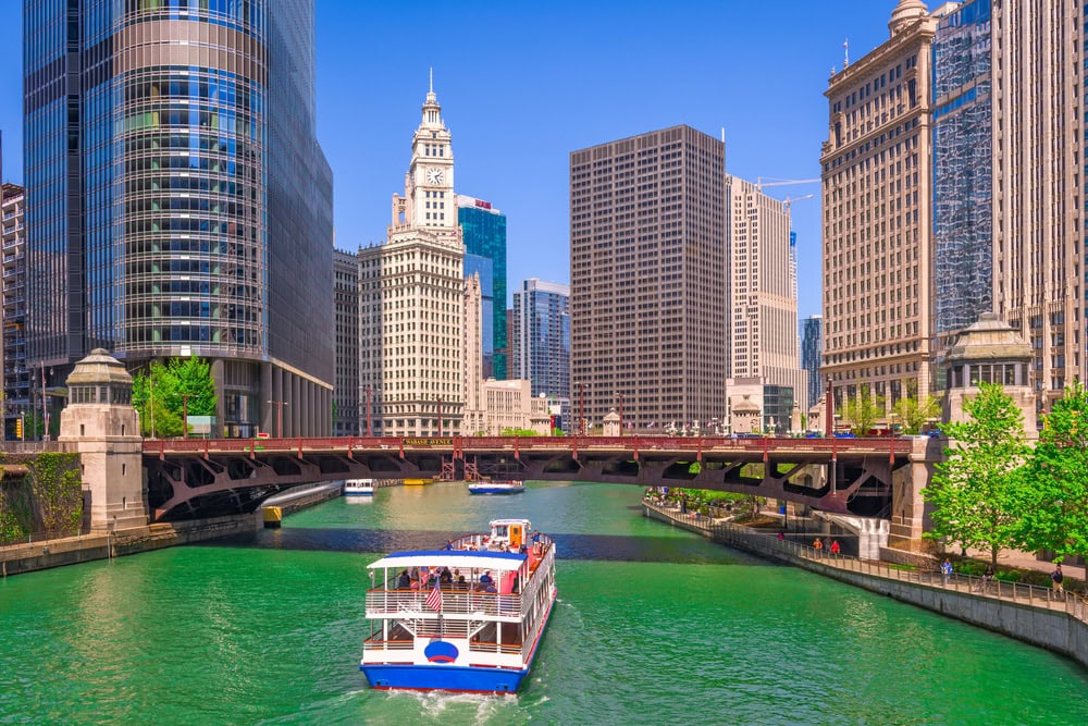 A tour boat travels along the Chicago River, surrounded by tall modern and historic buildings under a clear blue sky.