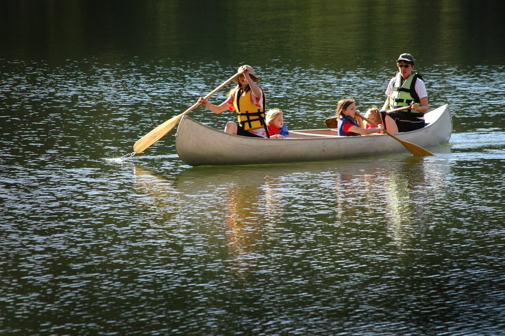 Four people wearing life jackets paddle a canoe on a calm lake, with sunlight reflecting off the water.
