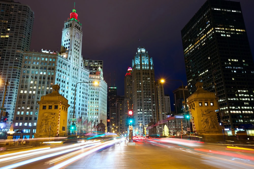 Nighttime cityscape with illuminated historic and modern buildings, light trails from moving vehicles, and stone bridge structures in downtown Chicago.