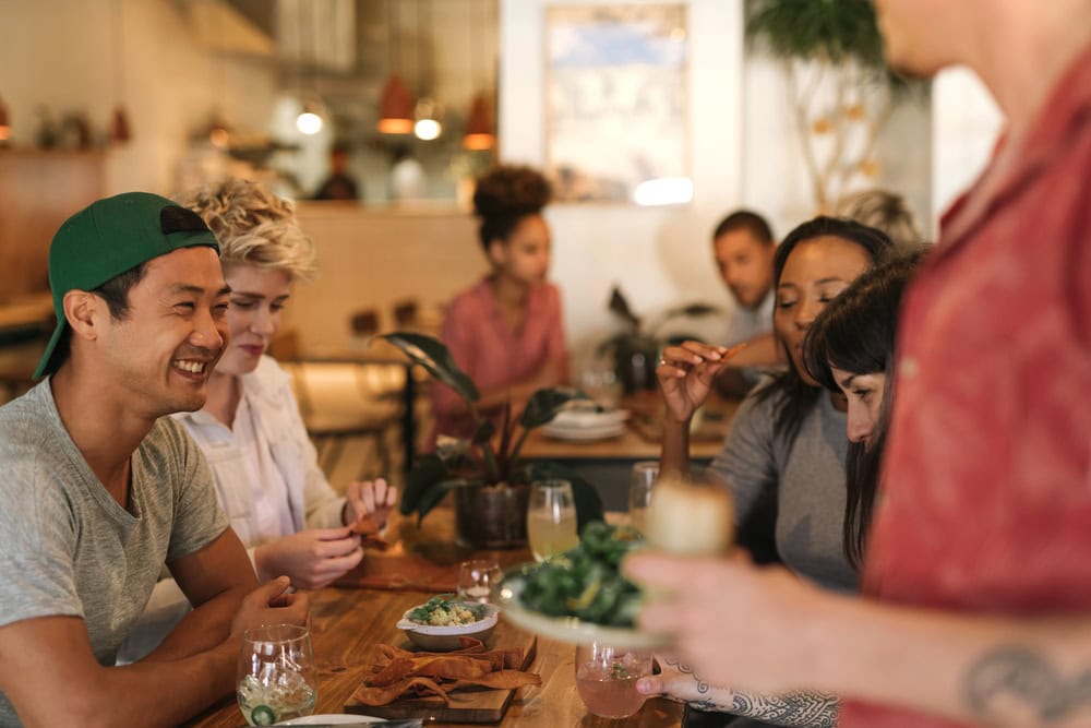 A group of people sit around a restaurant table eating and talking while a server brings food to the table.