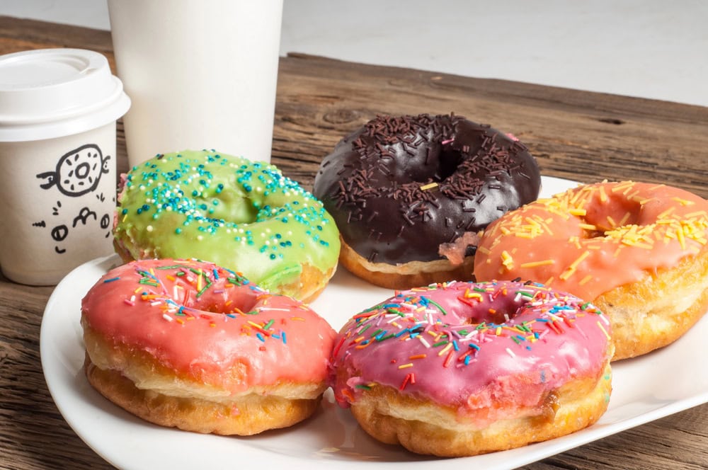 A plate of five frosted donuts with colorful sprinkles sits on a wooden table next to a white to-go coffee cup and a white cup.
