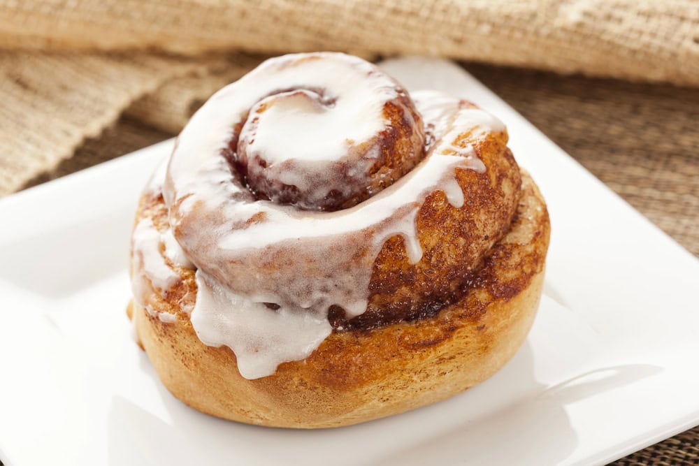 A cinnamon roll with white icing on top sits on a white square plate.