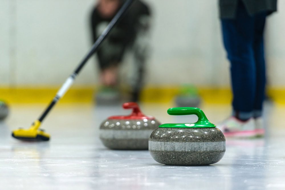Curling stones with red and green handles on ice; people playing in the background, one holding a yellow broom.