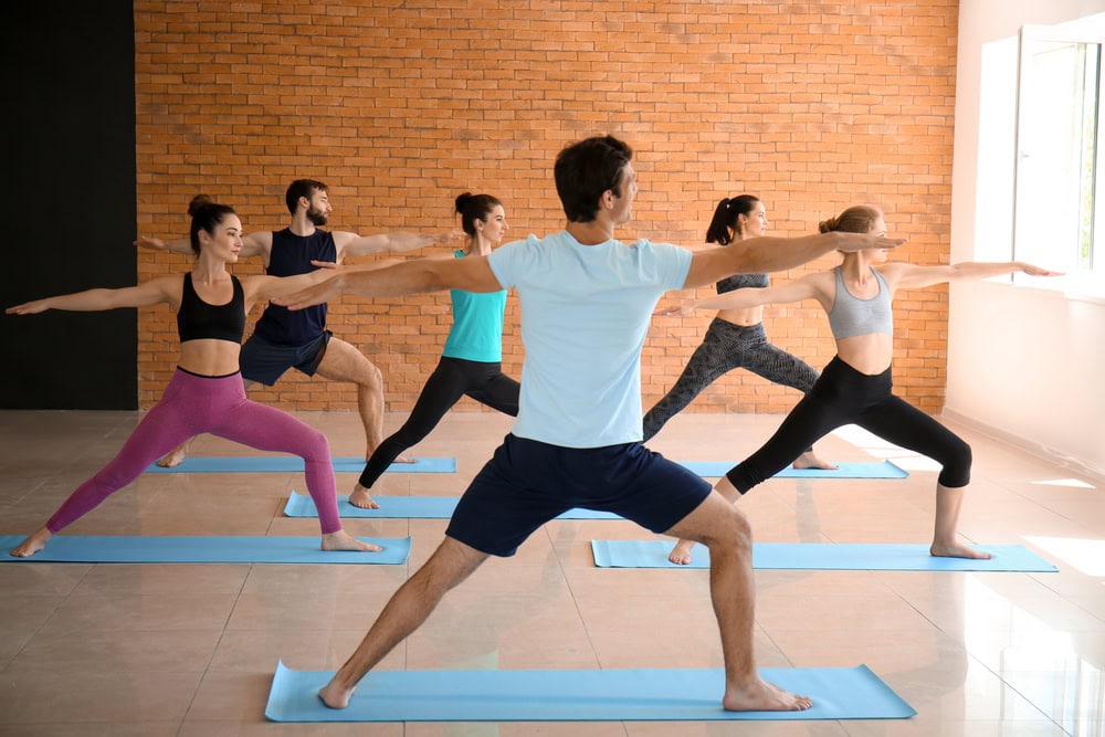 A group of people in athletic wear practice the Warrior II yoga pose on mats in a bright studio with a brick wall.