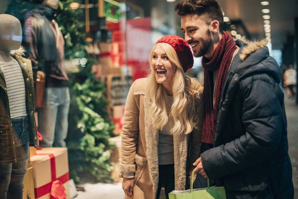 A smiling couple dressed in winter clothing stands outside a store, looking at a festive holiday window display with mannequins and gift boxes.