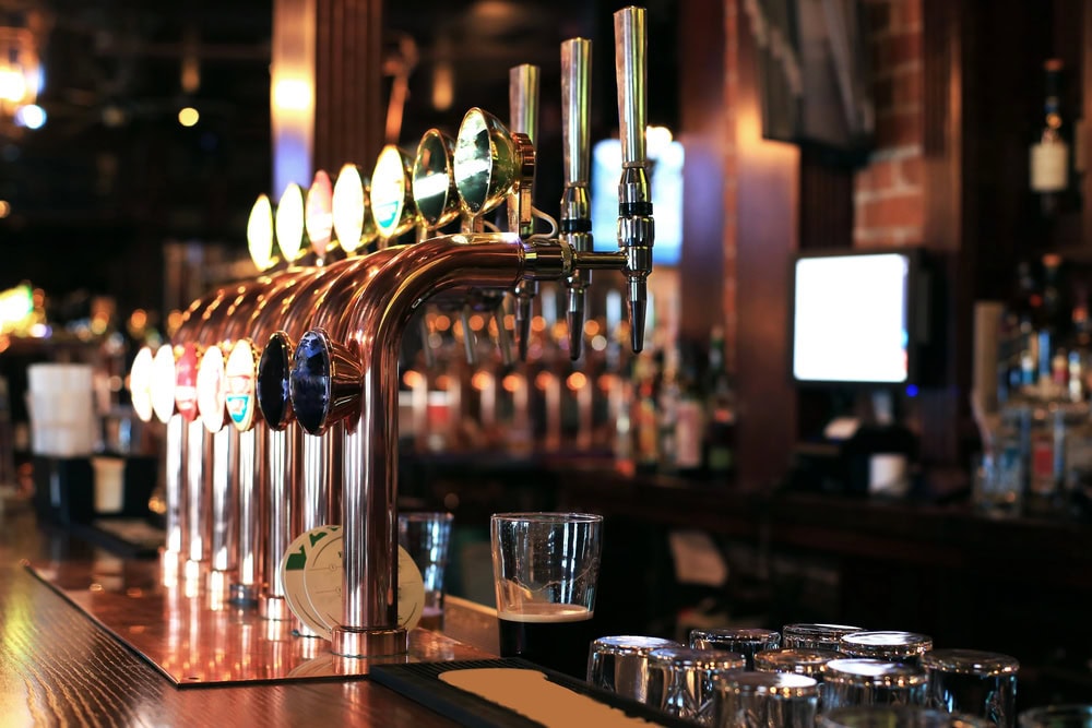 A row of beer taps is mounted on a bar counter with glasses and a partially filled pint glass in the foreground. The background shows shelves of bottles and a monitor.