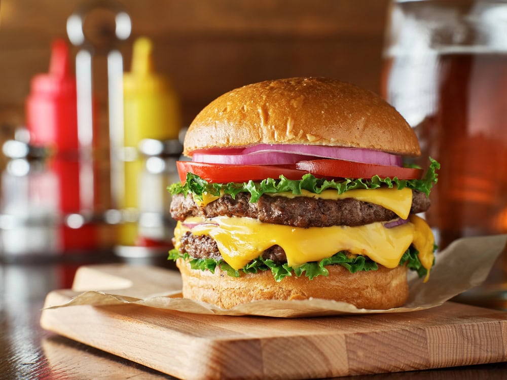 A cheeseburger with two beef patties, melted cheese, lettuce, tomato, red onion, and a bun sits on a wooden board, with condiments and a drink in the background.