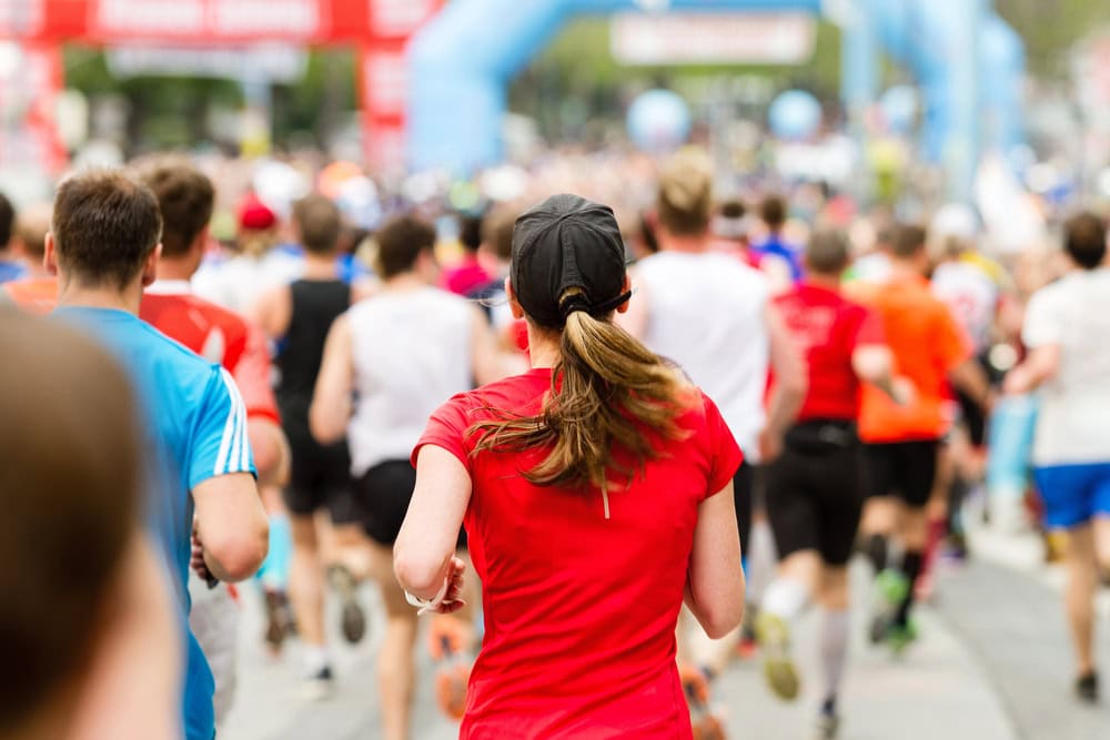A group of runners, seen from behind, participate in an outdoor race on a city street. The focus is on a woman in a red shirt and black cap.