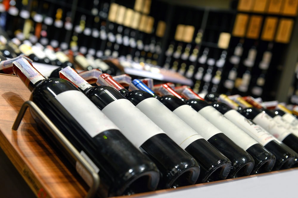 A row of wine bottles with blank labels is arranged on a wooden shelf in a wine store, with more bottles visible in the background.