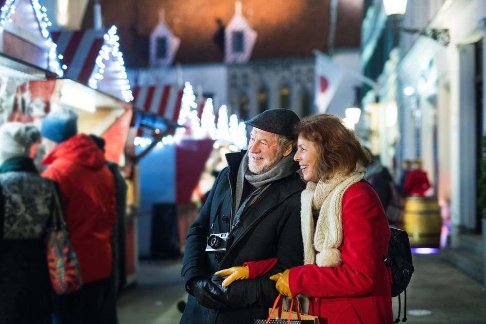 An older couple dressed in winter coats stand close together, smiling at an outdoor holiday market with festive lights and vendor stalls.