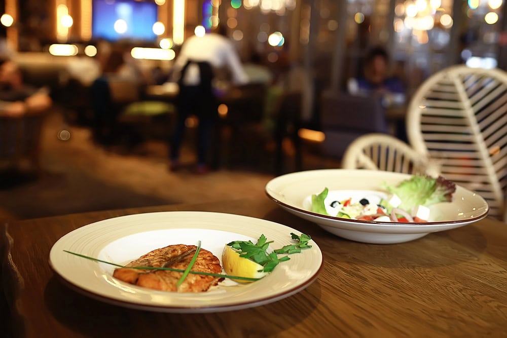 A plate of grilled fish with greens and lemon, and a bowl of salad on a wooden table in a restaurant with people seated in the background.