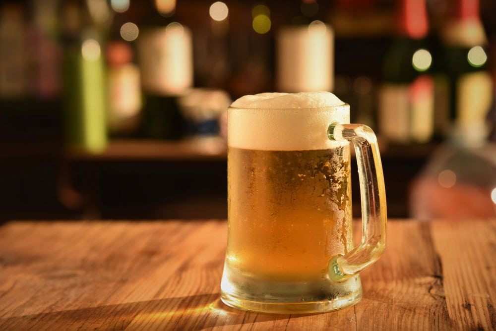 A frosty mug of beer with a foamy head sits on a wooden table, with blurred bottles and lights in the background.