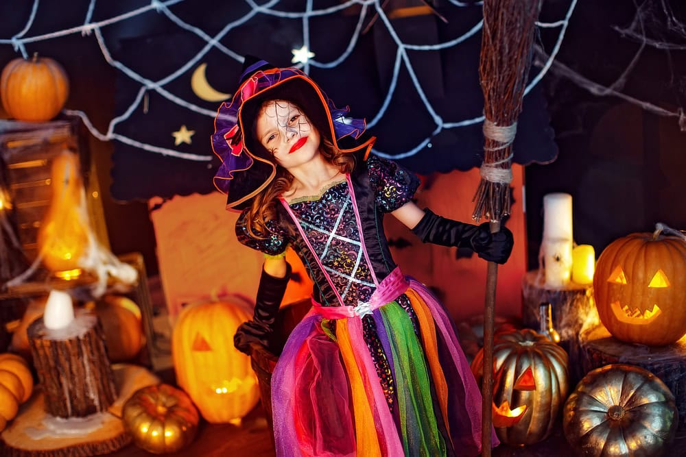 A child dressed as a colorful witch poses with a broomstick among carved pumpkins and Halloween decorations.