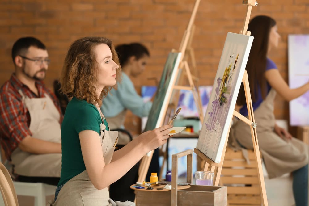 Several people wearing aprons sit at easels, painting on canvases in an art studio with a brick wall background.