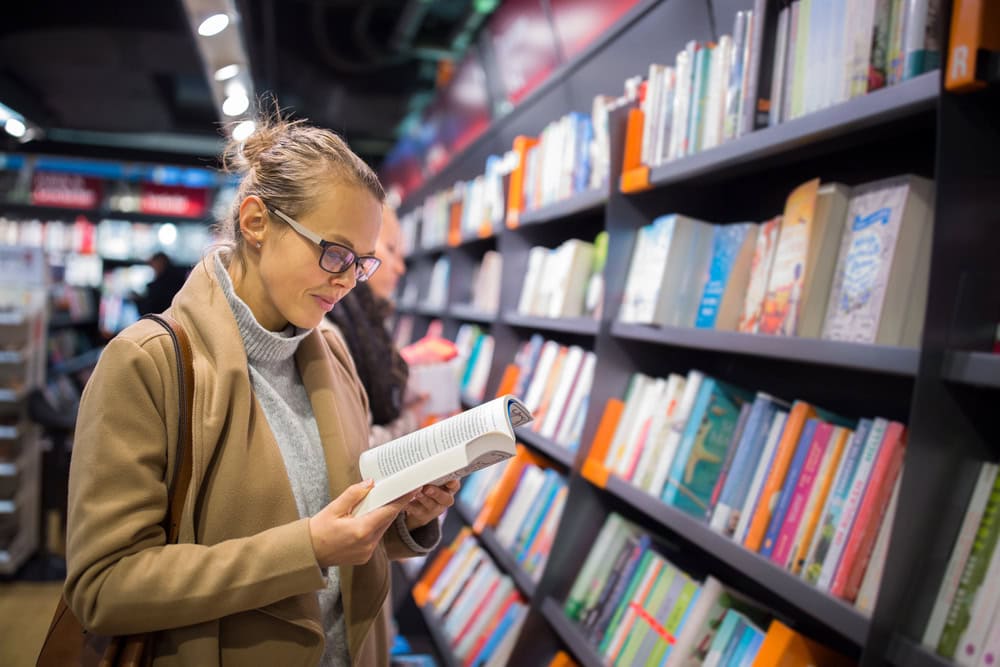 A woman wearing glasses and a tan coat reads a book while standing in front of bookshelves in a bookstore.