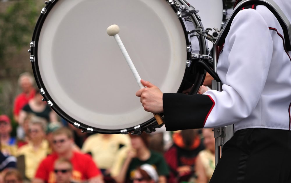 Person in a marching band uniform playing a bass drum with a mallet; blurred crowd seated in the background.