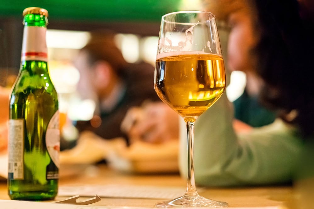 A glass of white wine and a green beer bottle are placed on a table, with blurred people and background in a bar or restaurant setting.