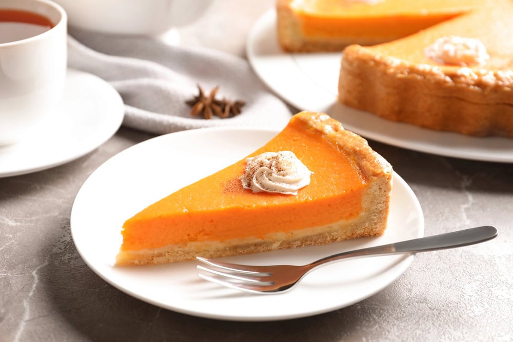A slice of pumpkin pie topped with whipped cream sits on a white plate with a fork, next to a cup of tea and a whole pie in the background.