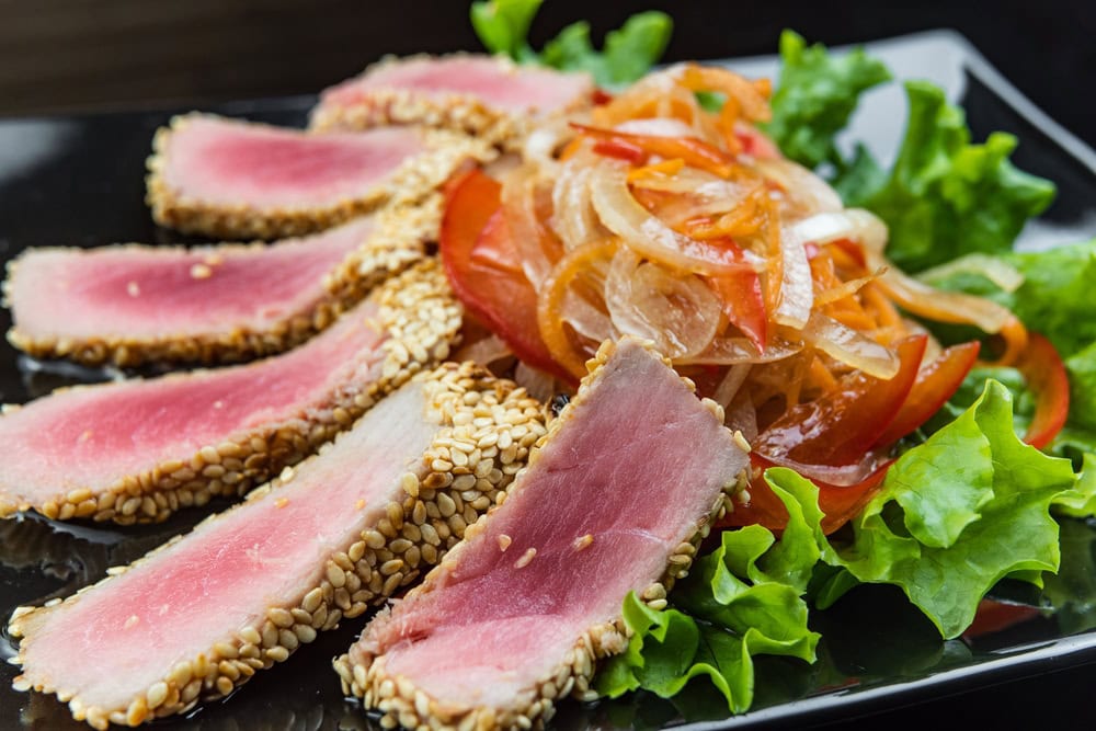 Slices of sesame-crusted seared tuna arranged on lettuce with a side of sautéed onions and red bell peppers on a black plate.