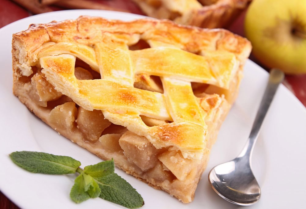 A slice of lattice-topped apple pie on a white plate with a spoon and mint garnish, with a whole apple in the background.