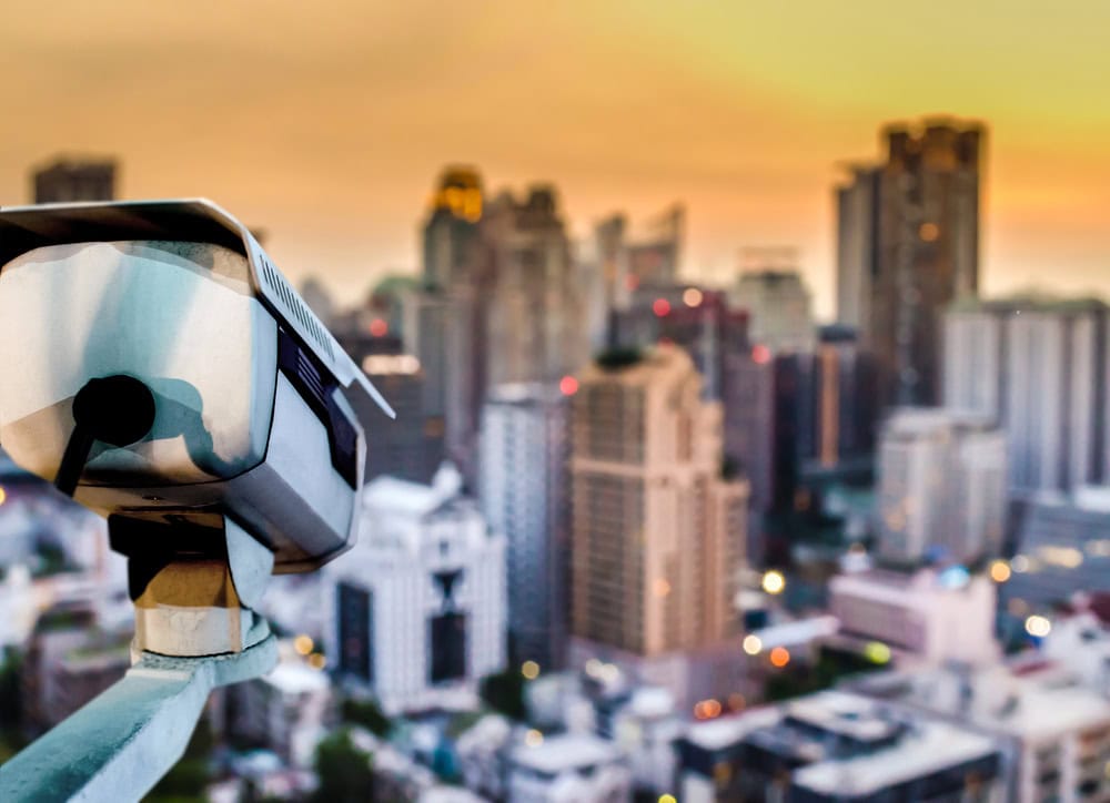 A security camera mounted in the foreground overlooks a cityscape with high-rise buildings at sunset.