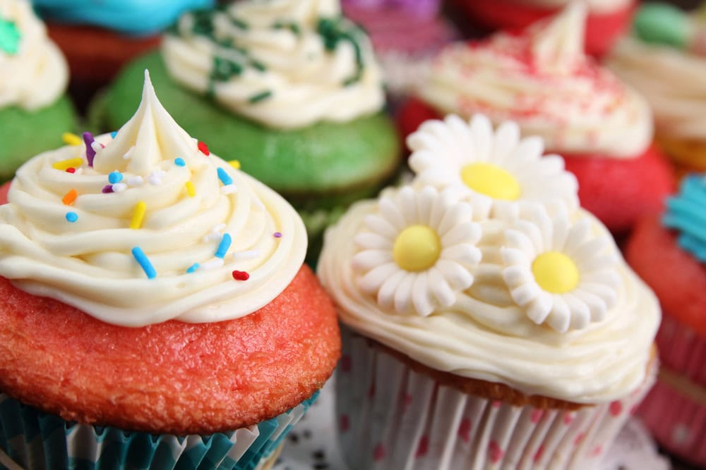 Several frosted cupcakes with colorful icing, sprinkles, and decorative flower toppers are arranged together on a surface.