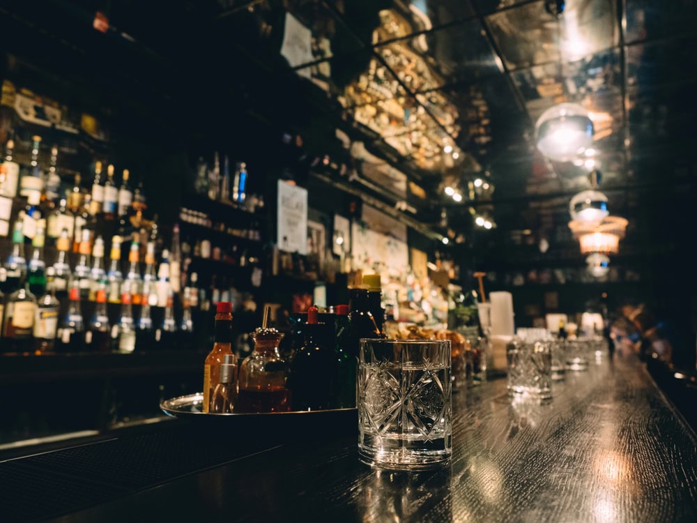 A dimly lit bar with glassware, bottles, and condiments on a wooden counter, and shelves of liquor in the background.