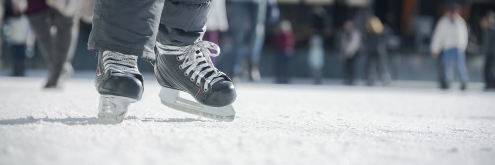 Close-up of a person wearing black ice skates on an outdoor ice rink, with people skating in the blurred background.