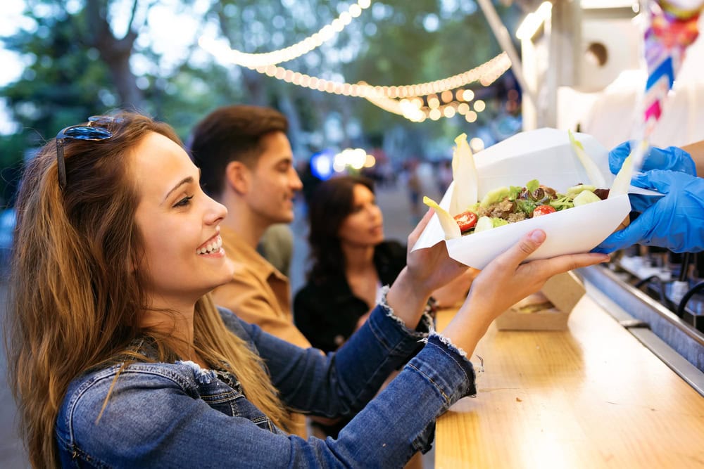 A woman smiles as she receives a container of food from a vendor at an outdoor food stall, with others waiting in line behind her.