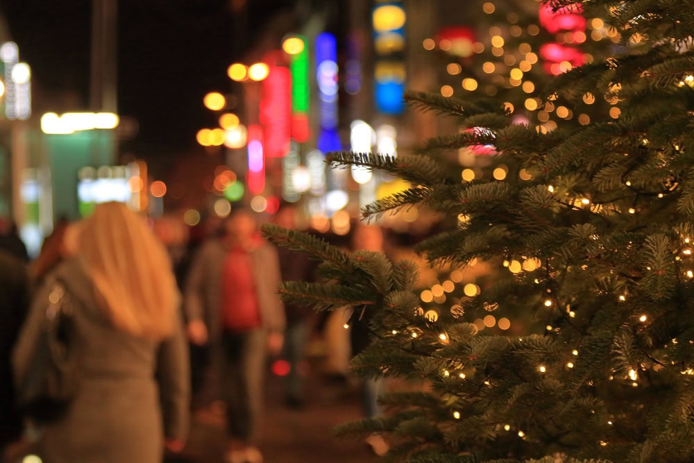A decorated Christmas tree with string lights is in focus on the right, while a busy city street with blurred people and colorful lights is in the background at night.