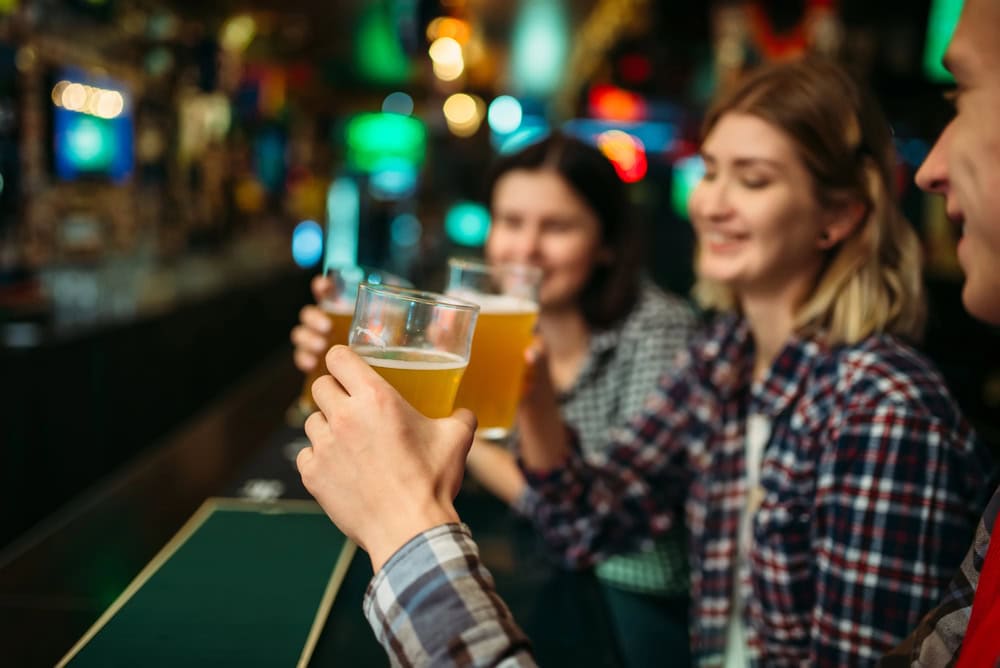 Three people sit at a bar holding pints of beer, smiling and raising their glasses in a toast.